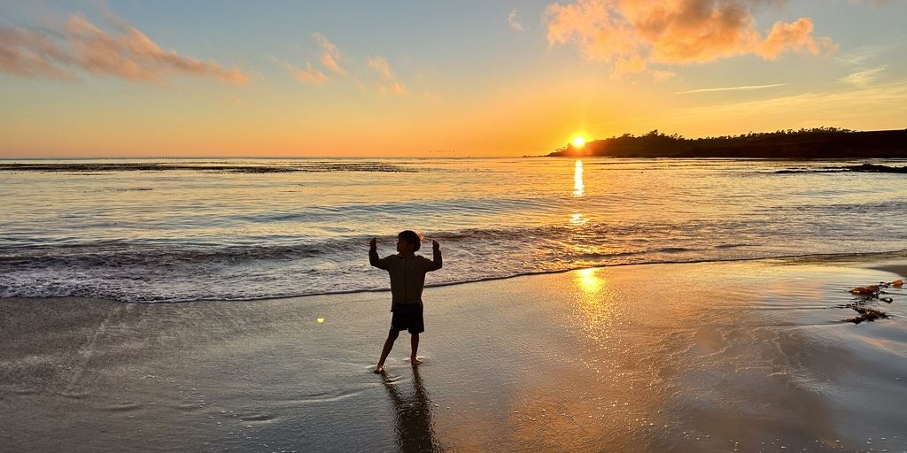 A child standing at the water's edge on Carmel Beach, silhouetted against the setting sun.